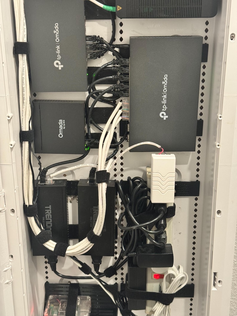 K&ouml;nnte beinhalten: Ein Netzwerkschrank mit mehreren TP-Link Omada- und Trendnet-Ger&auml;ten, die auf einer wei&szlig;en Platte montiert sind. Das Setup umfasst verschiedene schwarze Ethernet-Kabel, Netzteile und eine wei&szlig;e Steckdosenleiste, die mit schwarzen Klettb&auml;ndern befestigt sind.
