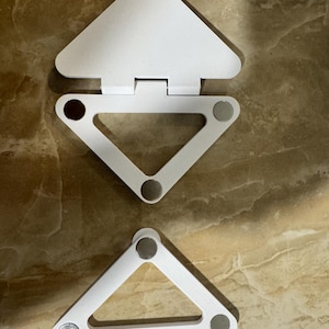 May include: Two white triangular folding stands made of plastic. The top stand features a hinged flap and three circular metallic magnets at the corners. The bottom stand is a simple triangular frame with three circular metallic magnets at the corners.