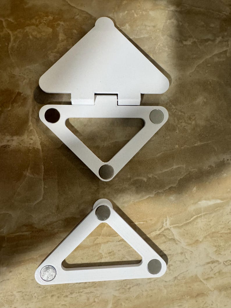 May include: Two white triangular folding stands made of plastic. The top stand features a hinged flap and three circular metallic magnets at the corners. The bottom stand is a simple triangular frame with three circular metallic magnets at the corners.
