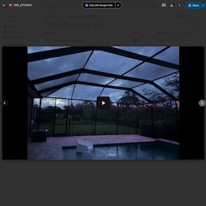 May include: A nighttime view of a pool area under a screened enclosure. The pool is surrounded by a brick patio and a black fence. String lights are visible in the background, and the sky is overcast.