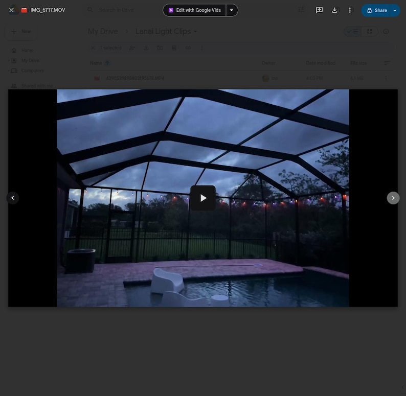 May include: A nighttime view of a pool area under a screened enclosure. The pool is surrounded by a brick patio and a black fence. String lights are visible in the background, and the sky is overcast.