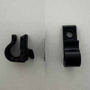 Könnte beinhalten: Zwei schwarze Kunststoffclips. Ein Clip hat eine gebogene Hakenform, der andere eine rechteckige Form mit einem Loch zur Montage. Beide Clips dienen zum Halten oder Sichern von Objekten.