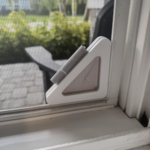 May include: A white triangular window screen corner reinforcement piece attached to the bottom corner of a window screen mesh. The plastic frame component features a hollow center and is secured against the white window frame and screen.