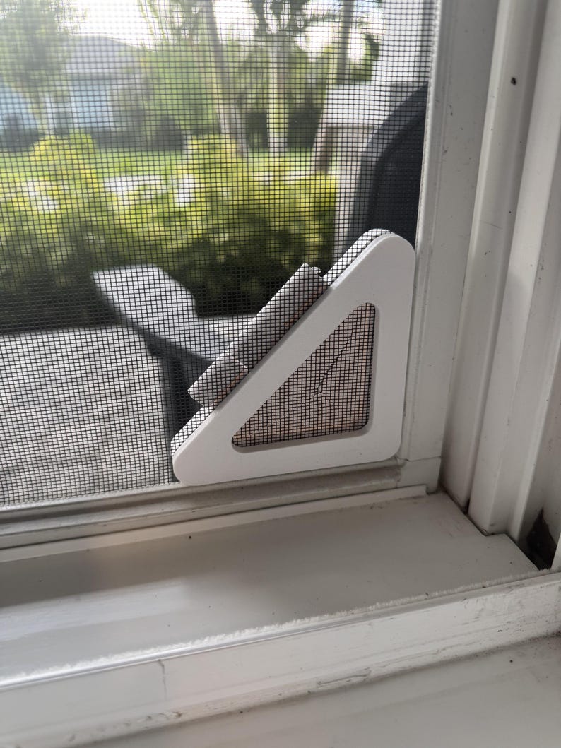 May include: A white triangular window screen corner reinforcement piece attached to the bottom corner of a window screen mesh. The plastic frame component features a hollow center and is secured against the white window frame and screen.