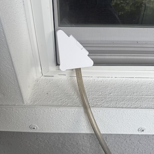 May include: A white, triangular-shaped plastic window vent adapter plate installed in a window frame. A clear, flexible plastic hose is attached to the bottom of the adapter, extending downwards away from the window screen and frame assembly.