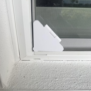 May include: A white triangular plastic corner clip attached to the bottom corner of a window screen frame. The clip features a hinged design and is positioned against the white window frame and the dark gray mesh screen material.