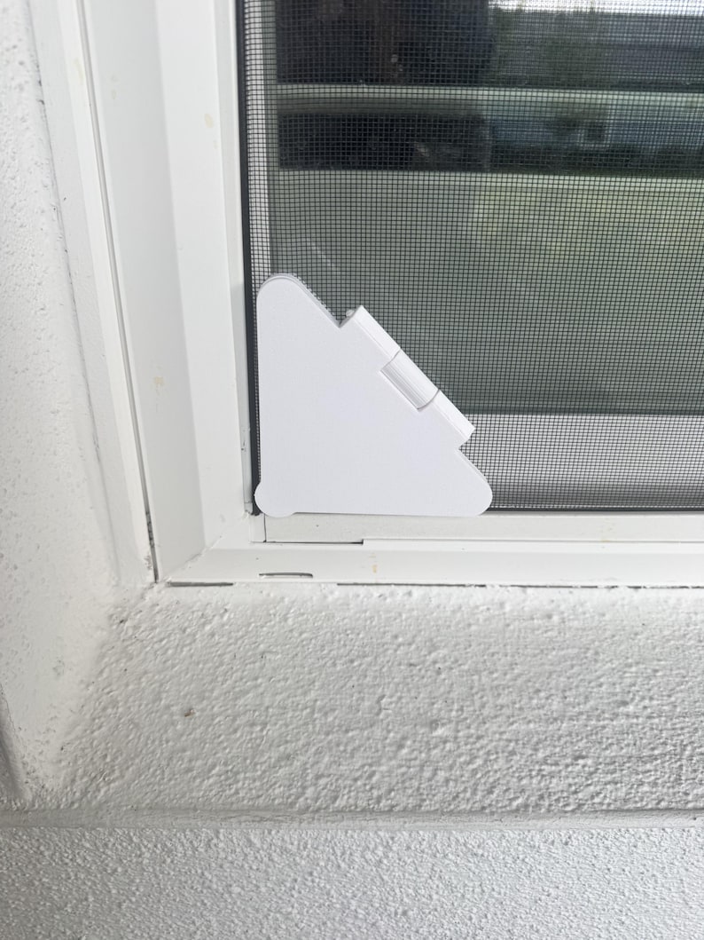 May include: A white triangular plastic corner clip attached to the bottom corner of a window screen frame. The clip features a hinged design and is positioned against the white window frame and the dark gray mesh screen material.