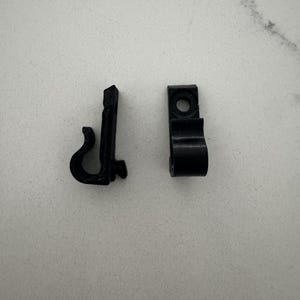 Puede incluir: Dos pequeños clips de plástico negro. Uno tiene forma de gancho curvo y el otro tiene un cuerpo rectangular con un agujero y una sección curva. Estos clips probablemente se utilizan para colgar o asegurar objetos.