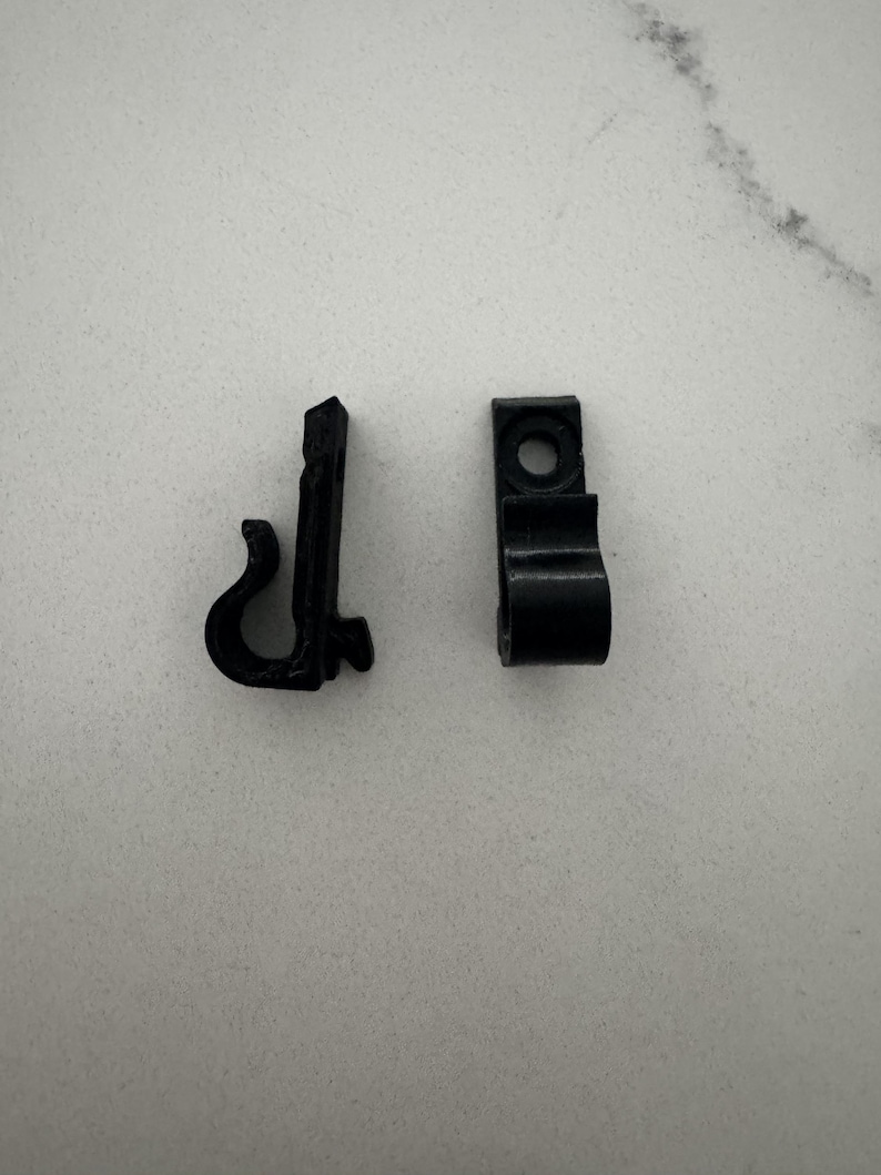 May include: Two small, black plastic clips. One clip has a curved hook shape, and the other has a rectangular body with a hole and a curved section. These clips are likely used for hanging or securing items.