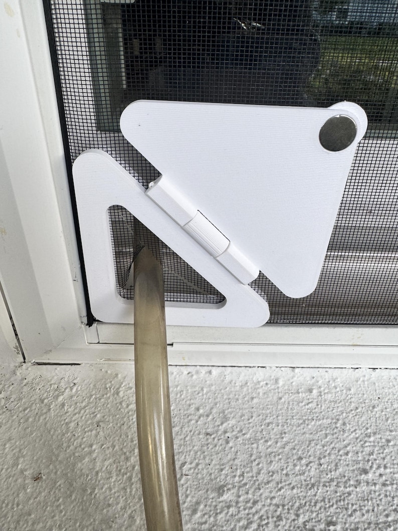 May include: A white plastic triangular pet door insert installed into a black mesh screen door. The device features a hinged flap mechanism and a circular silver-colored magnetic closure at the top corner, with a clear plastic hose passing through.