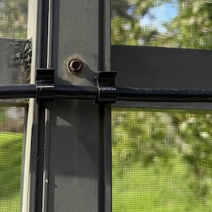 May include: Two black plastic cable clips are attached to a vertical gray metal frame. A thin black cable runs horizontally through the center of both clips. A single rusted metal bolt is visible on the gray frame above the cable clips.