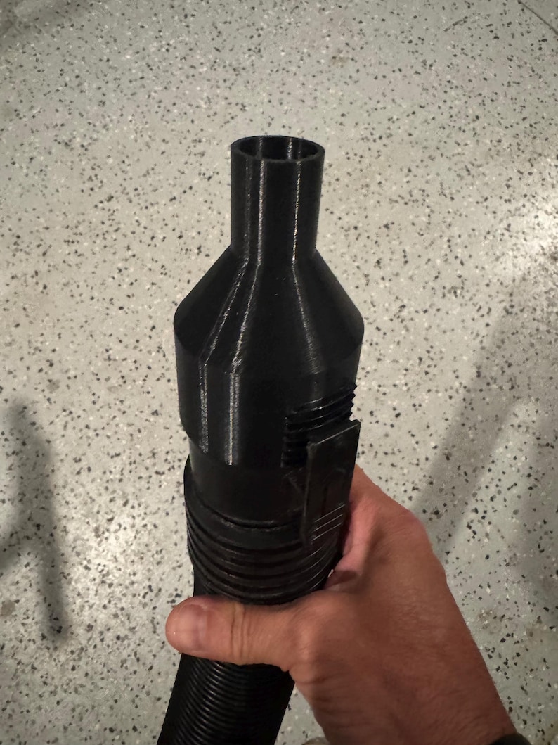 May include: A black, 3D-printed object with a cylindrical, ribbed handle and a flared top. The object has a rectangular clip and a series of ridges. The item is held in a hand.