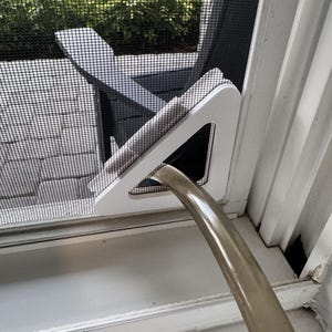 May include: A white triangular window screen insert featuring a central opening designed to accommodate a flexible, translucent hose. The insert is securely positioned within the frame of a black mesh screen door, allowing the hose to pass through.
