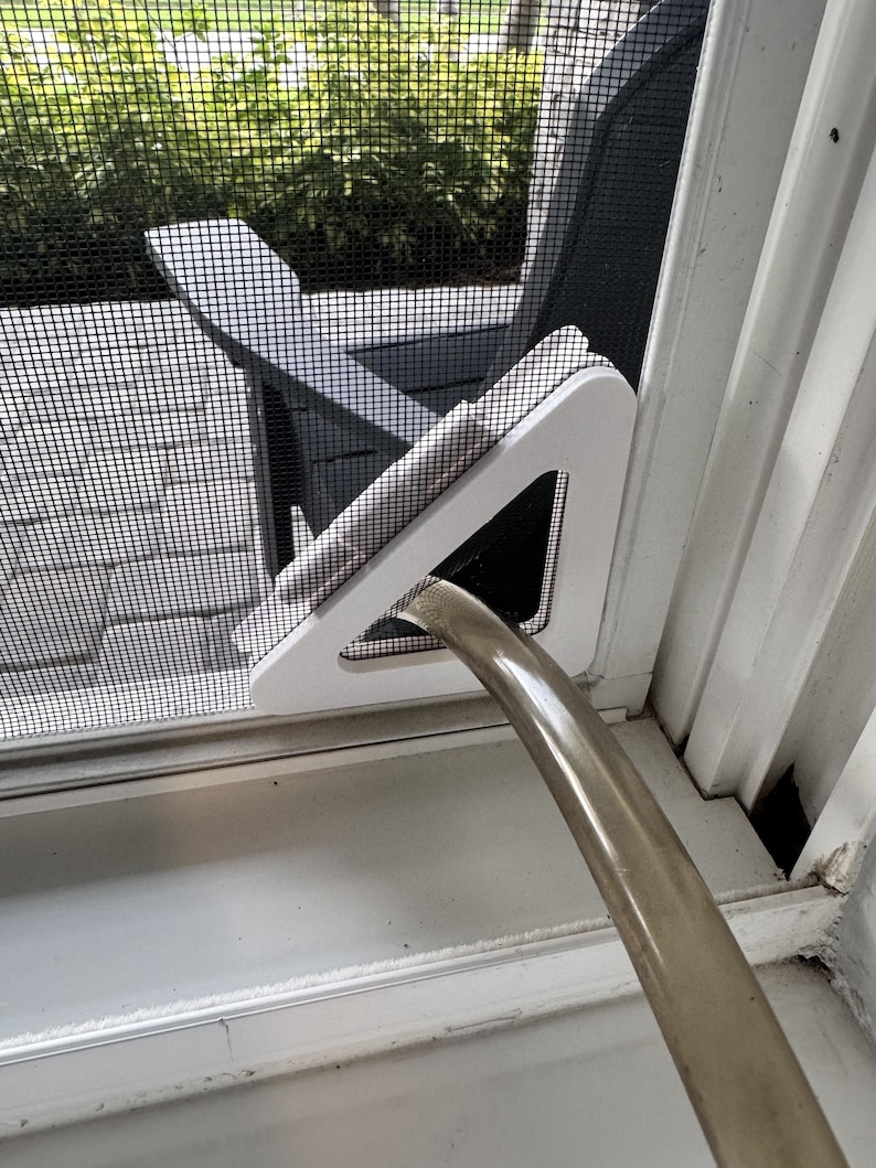May include: A white triangular window screen insert featuring a central opening designed to accommodate a flexible, translucent hose. The insert is securely positioned within the frame of a black mesh screen door, allowing the hose to pass through.