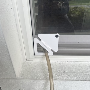 May include: A white triangular window screen adapter plate with a hinged design and a circular black fastener. The device is installed in the corner of a window frame, featuring a central circular opening that holds a flexible, translucent hose.