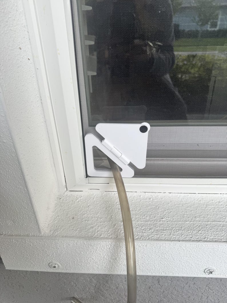 May include: A white triangular window screen adapter plate with a hinged design and a circular black fastener. The device is installed in the corner of a window frame, featuring a central circular opening that holds a flexible, translucent hose.