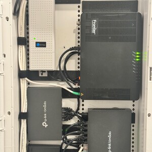 K&ouml;nnte beinhalten: Ein wei&szlig;es Wandgeh&auml;use f&uuml;r Netzwerktechnik mit einem wei&szlig;en Frontier ONT-Ger&auml;t, einem schwarzen Frontier-Gateway und drei schwarzen TP-Link Omada-Netzwerkger&auml;ten. Mehrere schwarze und wei&szlig;e Ethernet-Kabel sind an die Anschl&uuml;sse angeschlossen.
