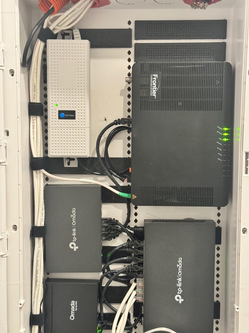 K&ouml;nnte beinhalten: Ein wei&szlig;es Wandgeh&auml;use f&uuml;r Netzwerktechnik mit einem wei&szlig;en Frontier ONT-Ger&auml;t, einem schwarzen Frontier-Gateway und drei schwarzen TP-Link Omada-Netzwerkger&auml;ten. Mehrere schwarze und wei&szlig;e Ethernet-Kabel sind an die Anschl&uuml;sse angeschlossen.