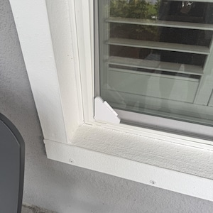 May include: A white plastic corner bracket installed on the bottom corner of a window frame. The bracket is triangular with a flat surface and is mounted against the white window casing and the glass pane, securing the screen frame in place.