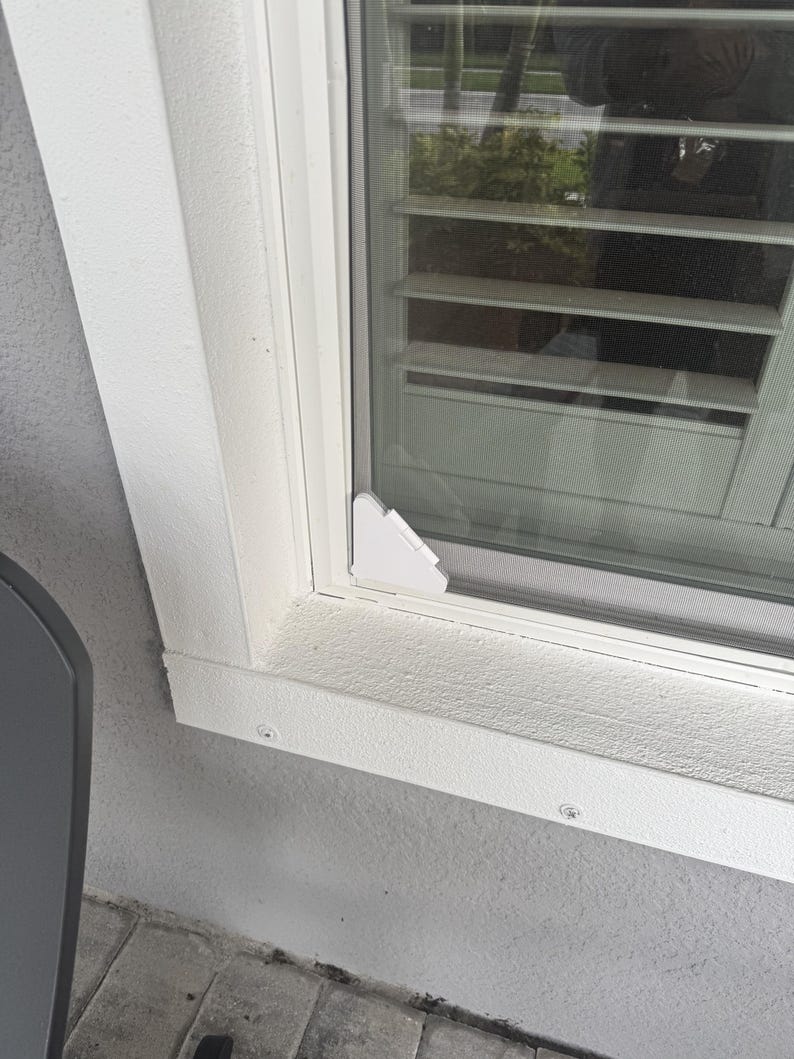 May include: A white plastic corner bracket installed on the bottom corner of a window frame. The bracket is triangular with a flat surface and is mounted against the white window casing and the glass pane, securing the screen frame in place.