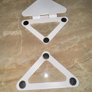 May include: Two white triangular stands with a smooth finish. Each stand features three circular black pads on the underside for stability. The top stand includes a hinged triangular flap attached to the base, while the bottom stand is a single frame.