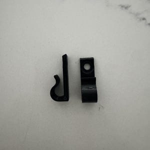 May include: Two small, black plastic clips. One is L-shaped with a curved hook. The other is rectangular with a circular hole. Both clips are on a white, speckled surface.