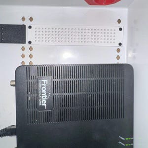 K&ouml;nnte beinhalten: Ein schwarzer Frontier Communications Netzwerk-Router mit vertikalen L&uuml;ftungsschlitzen und gr&uuml;nen Statusanzeigen mit der Beschriftung Power, Optical, WAN, Update, Alarm und POTS, montiert in einem wei&szlig;en Geh&auml;use mit wei&szlig;er Lochplatte.