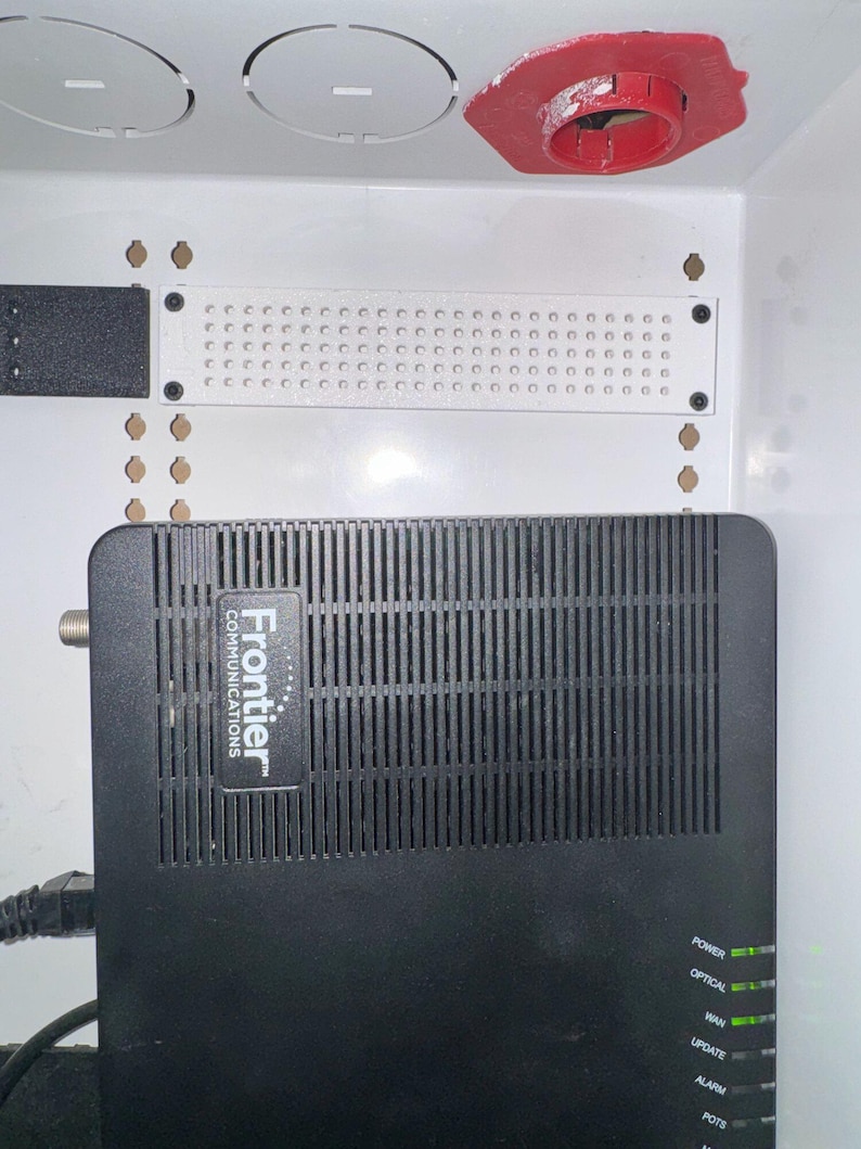 K&ouml;nnte beinhalten: Ein schwarzer Frontier Communications Netzwerk-Router mit vertikalen L&uuml;ftungsschlitzen und gr&uuml;nen Statusanzeigen mit der Beschriftung Power, Optical, WAN, Update, Alarm und POTS, montiert in einem wei&szlig;en Geh&auml;use mit wei&szlig;er Lochplatte.