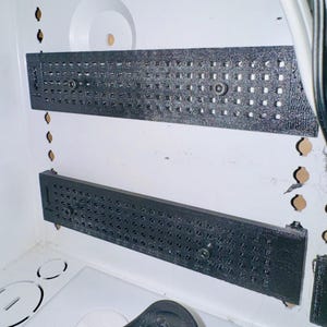 Puede incluir: Dos soportes metálicos negros, rectangulares y perforados, están montados dentro de una carcasa blanca. Los soportes están asegurados con tornillos. Un enchufe eléctrico negro es visible en la parte inferior de la imagen.