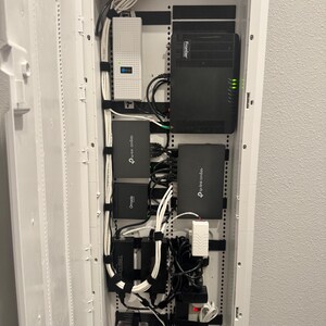 K&ouml;nnte beinhalten: Ein wei&szlig;es, an der Wand montiertes Mediengeh&auml;use mit verschiedenen Netzwerkger&auml;ten, darunter ein schwarzer Frontier-Router, mehrere TP-Link Omada-Ger&auml;te, ein wei&szlig;es Netzteil und organisierte wei&szlig;e Ethernet-Kabel, die mit schwarzen Klettb&auml;ndern befestigt sind.