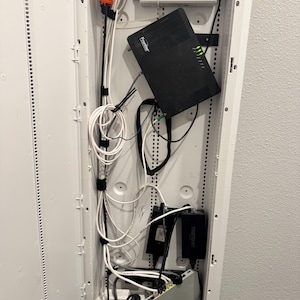 K&ouml;nnte beinhalten: Ein wei&szlig;er, an der Wand montierter Medienverteiler mit einem schwarzen Frontier-Router mit gr&uuml;nen Kontrollleuchten, einem wei&szlig;en Netzteil, zwei schwarzen Trendnet-Adaptern und einem silbernen Netzwerk-Switch, verbunden durch wei&szlig;e und schwarze Kabel.