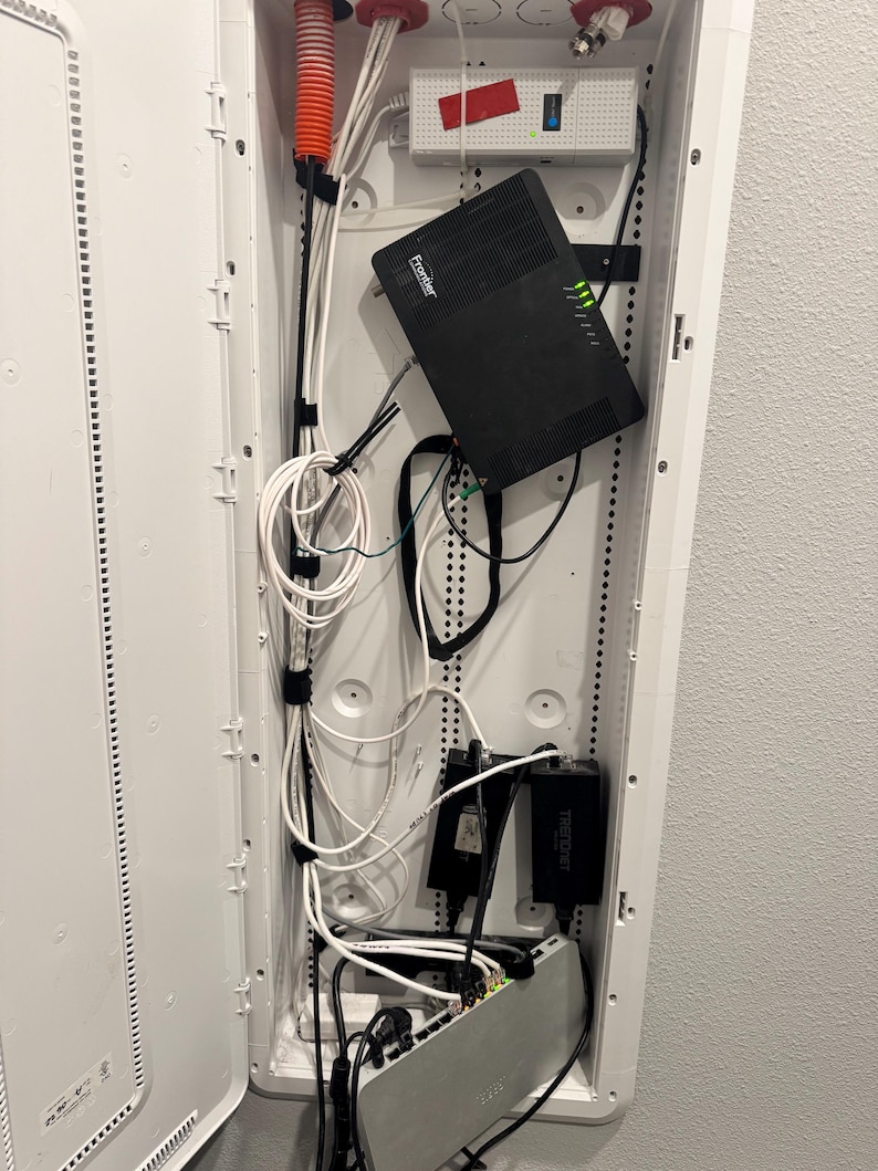 K&ouml;nnte beinhalten: Ein wei&szlig;er, an der Wand montierter Medienverteiler mit einem schwarzen Frontier-Router mit gr&uuml;nen Kontrollleuchten, einem wei&szlig;en Netzteil, zwei schwarzen Trendnet-Adaptern und einem silbernen Netzwerk-Switch, verbunden durch wei&szlig;e und schwarze Kabel.