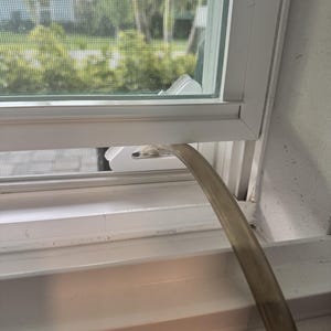 May include: A white plastic window vent adapter plate installed in a window frame, featuring a circular opening connected to a flexible, translucent grey hose that extends outward, facilitating airflow through the screen mesh of the window.
