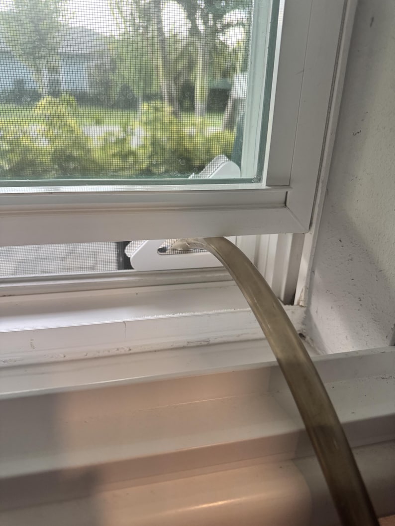 May include: A white plastic window vent adapter plate installed in a window frame, featuring a circular opening connected to a flexible, translucent grey hose that extends outward, facilitating airflow through the screen mesh of the window.