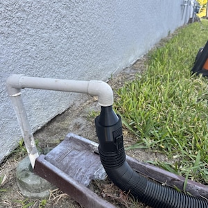 May include: A grey PVC pipe connected to a black corrugated hose, leading to a brown metal gutter. The gutter is placed on the ground near a grassy area and a light grey stucco wall.
