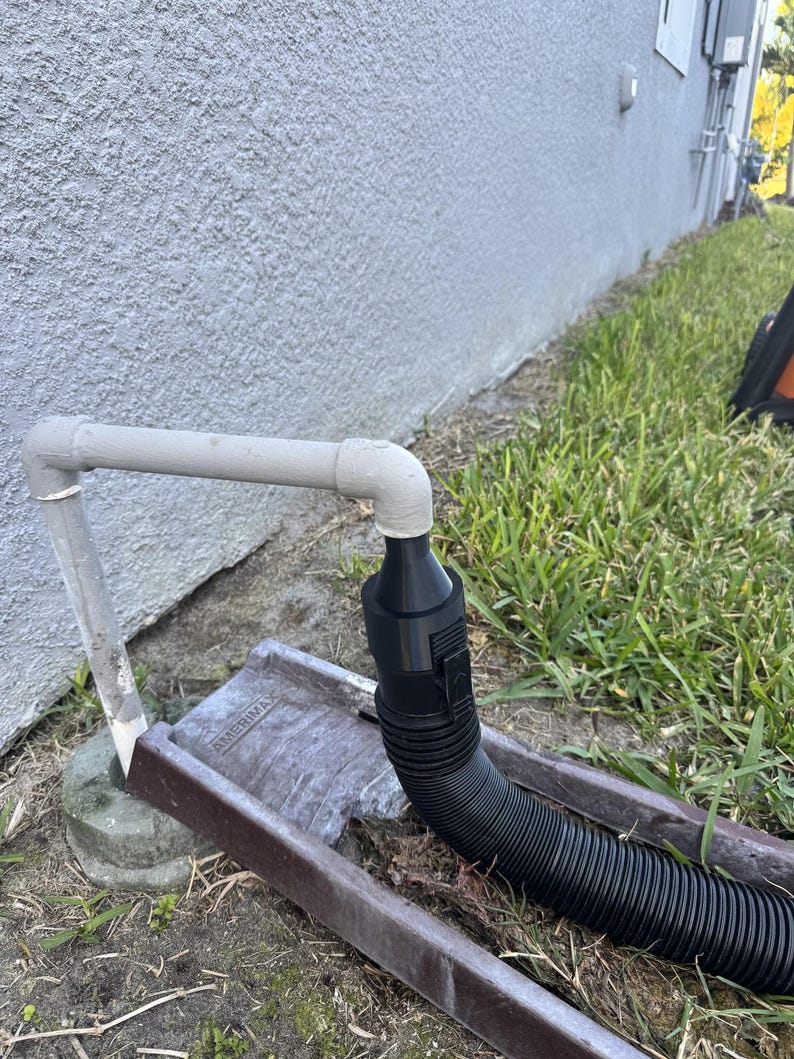 May include: A grey PVC pipe connected to a black corrugated hose, leading to a brown metal gutter. The gutter is placed on the ground near a grassy area and a light grey stucco wall.