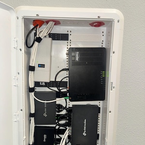 K&ouml;nnte beinhalten: Ein wei&szlig;es, an der Wand montiertes Mediengeh&auml;use mit einem schwarzen Frontier-Router, zwei schwarzen TP-Link Omada-Netzwerkger&auml;ten und einem wei&szlig;en Netzteil. Mehrere wei&szlig;e Ethernet-Kabel sind mit schwarzen Klettb&auml;ndern geordnet.