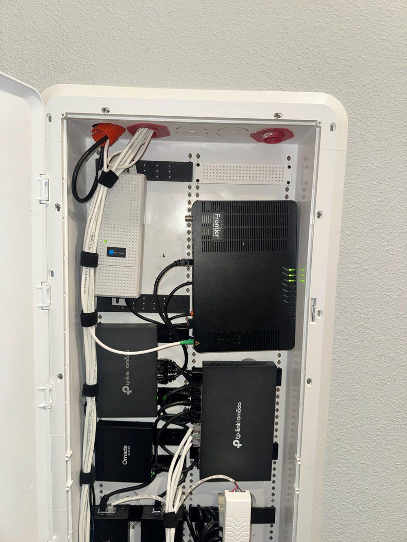 K&ouml;nnte beinhalten: Ein wei&szlig;es, an der Wand montiertes Mediengeh&auml;use mit einem schwarzen Frontier-Router, zwei schwarzen TP-Link Omada-Netzwerkger&auml;ten und einem wei&szlig;en Netzteil. Mehrere wei&szlig;e Ethernet-Kabel sind mit schwarzen Klettb&auml;ndern geordnet.