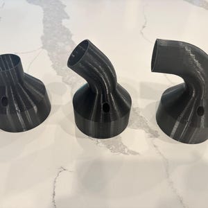 Puede incluir: Tres boquillas negras impresas en 3D con un diseño curvo. Cada boquilla tiene una base ancha, un orificio central y una salida más estrecha e inclinada. Los objetos están dispuestos sobre una superficie blanca estampada.