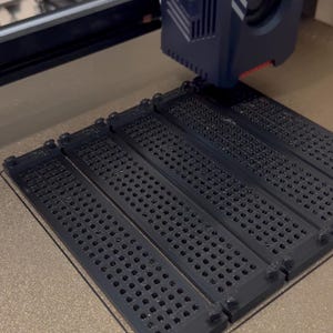 K&ouml;nnte beinhalten: Ein dunkelblauer 3D-Drucker-Extruderkopf befindet sich &uuml;ber einer Druckplatte und druckt vier identische schwarze rechteckige Bauteile. Jedes Bauteil weist ein Gitter aus kleinen runden L&ouml;chern und erhabenen runden Noppen an den Ecken auf.