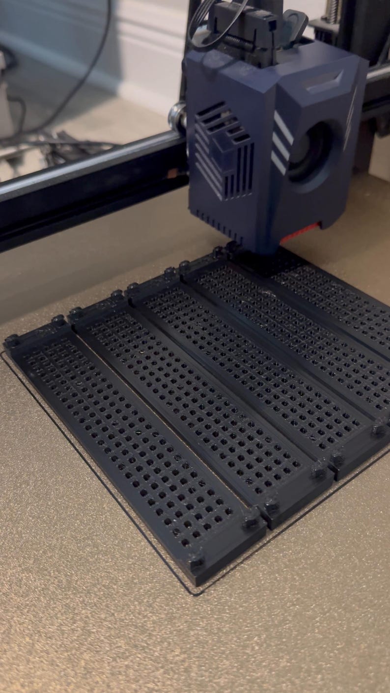K&ouml;nnte beinhalten: Ein dunkelblauer 3D-Drucker-Extruderkopf befindet sich &uuml;ber einer Druckplatte und druckt vier identische schwarze rechteckige Bauteile. Jedes Bauteil weist ein Gitter aus kleinen runden L&ouml;chern und erhabenen runden Noppen an den Ecken auf.