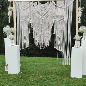 May include: A macrame wedding backdrop with a geometric design, hanging from a wooden frame. White pedestals hold floral arrangements, and the scene is set on a grassy lawn. The backdrop features intricate knotting and fringe.
