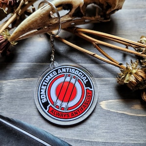 Puede incluir: Un llavero redondo de acrílico transparente con un diseño en blanco y negro. El diseño presenta un círculo rojo con tres flechas apuntando hacia él. El texto "Sometimes Antisocial Always Anti-Fascist" está escrito alrededor del círculo.