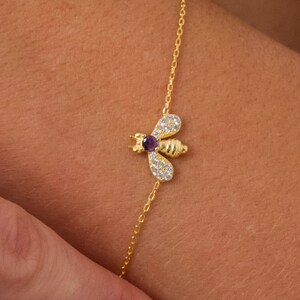 May include: A gold chain bracelet with a small bee charm. The bee has a purple gemstone for its body and clear gemstones for its wings.