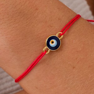 Könnte beinhalten: Rotes Armband mit einem goldfarbenen bösen Blick-Anhänger. Der Anhänger ist blau mit einem weißen und gelben Auge.