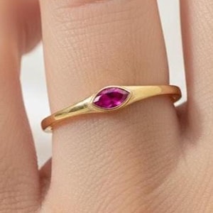 May include: A gold ring featuring a marquise-cut, ruby-coloured gemstone. The ring's band is a thin, polished gold. The gemstone is set horizontally in the centre of the ring, creating a sleek design. The ring is shown on a finger.