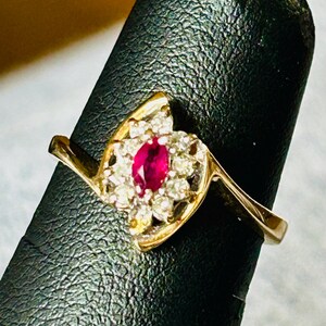 May include: A gold ring with a red ruby gemstone surrounded by smaller white diamonds. The ring has a unique, curved design.