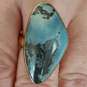 May include: A gold-toned ring featuring a large, elongated, teardrop-shaped stone. The stone has a marbled appearance with shades of blue and black. The ring is set in a simple, polished gold band.