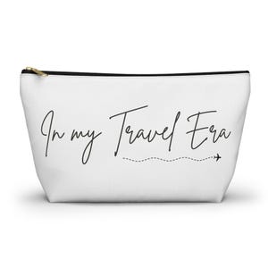 May include: White zippered pouch with the text "In my Travel Era" in black cursive font. A small airplane icon is next to a dotted line.
