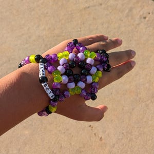 May include: A star-shaped beaded bracelet in shades of purple, green, black, and white, spelling out "SAVA BOYS" on one strand. The bracelet is made of small, colorful beads and is worn on a hand.